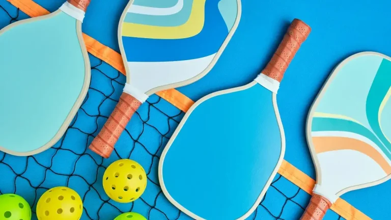 Colorful pickleball paddles and balls laid out on a court next to a net, representing how to choose the right pickleball paddle for your game.